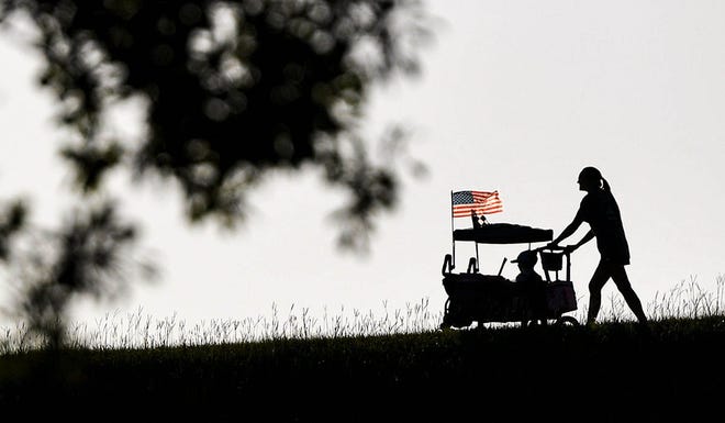 A woman pushes her child in a cart with a U.S. flag waving from the top, walking along the dike along the Seneca River of Lake Hartwell looking for a good viewing spot before the Clemson Light the Lake Fireworks event in Clemson, S.C. Wednesday, July 3, 2024.